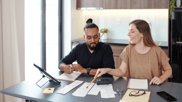 A couple sitting at a kitchen table, reviewing receipts and financial documents while budgeting. The man, with a beard and tied-up hair, is taking notes in a notebook, while the woman, wearing a brown t-shirt, is smiling and pointing at receipts with a pencil. A calculator, glasses, and a tablet stand are also on the table.