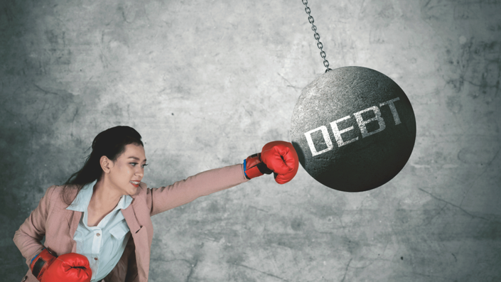 A determined woman wearing red boxing gloves punches a large wrecking ball labeled "DEBT," symbolizing the power of Debt Management in overcoming financial struggles.
