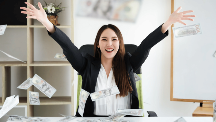A joyful woman in a business suit sits at a desk with her arms raised, celebrating as money floats around her, symbolizing financial success and freedom.