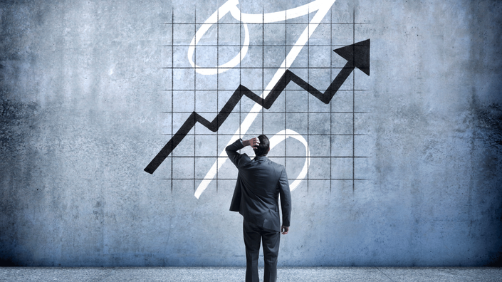 A businessman in a suit stands in front of a large wall displaying a grid with a percentage symbol and an upward-trending arrow. He scratches his head while looking at the graph, symbolizing rising interest rates and financial uncertainty.