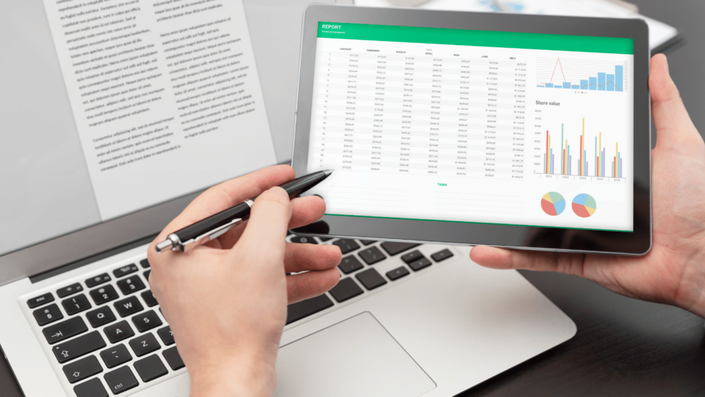 A person holding a tablet displaying financial reports with spreadsheets, charts, and graphs while working on a laptop. This image symbolizes strategic financial management and creating a Realistic budget for long-term financial stability.