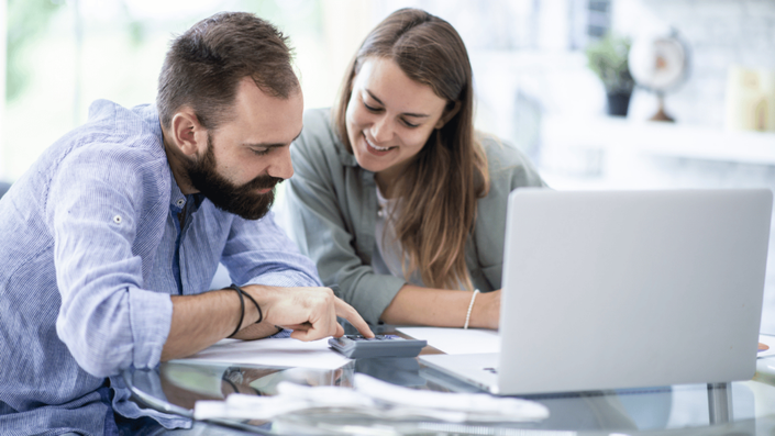 A couple sitting at a glass table, smiling and working on their personal finance. The man is using a calculator while the woman looks on, appearing engaged in the financial discussion. A laptop is open on the table, indicating they may be managing their budget or expenses.