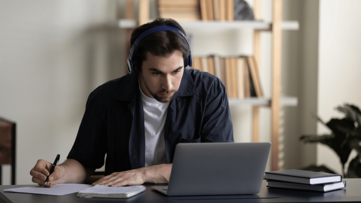 Focused young man wearing headphones, taking notes while studying personal finance online on his laptop at a desk with books in the background.