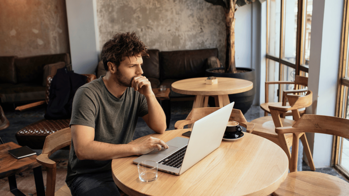 A young man with a beard and casual attire sits in a modern café, focused on his laptop. He appears deep in thought, with a coffee cup and water glass on the wooden table. The scene represents remote work flexibility and the freelancer lifestyle.